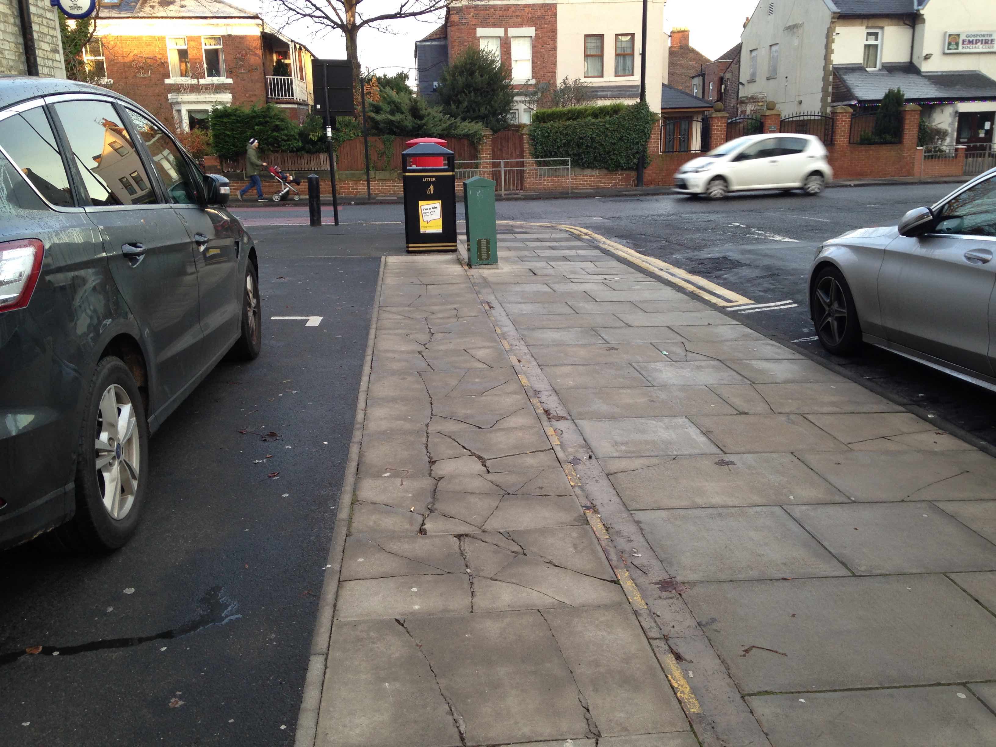 Photograph of broken and uneven paving stones between the parking spaces, the pavement and the road
